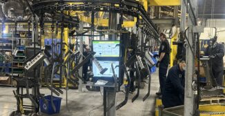 Operators working alongside a powder coating conveyor line with a job display screen on the shop floor
