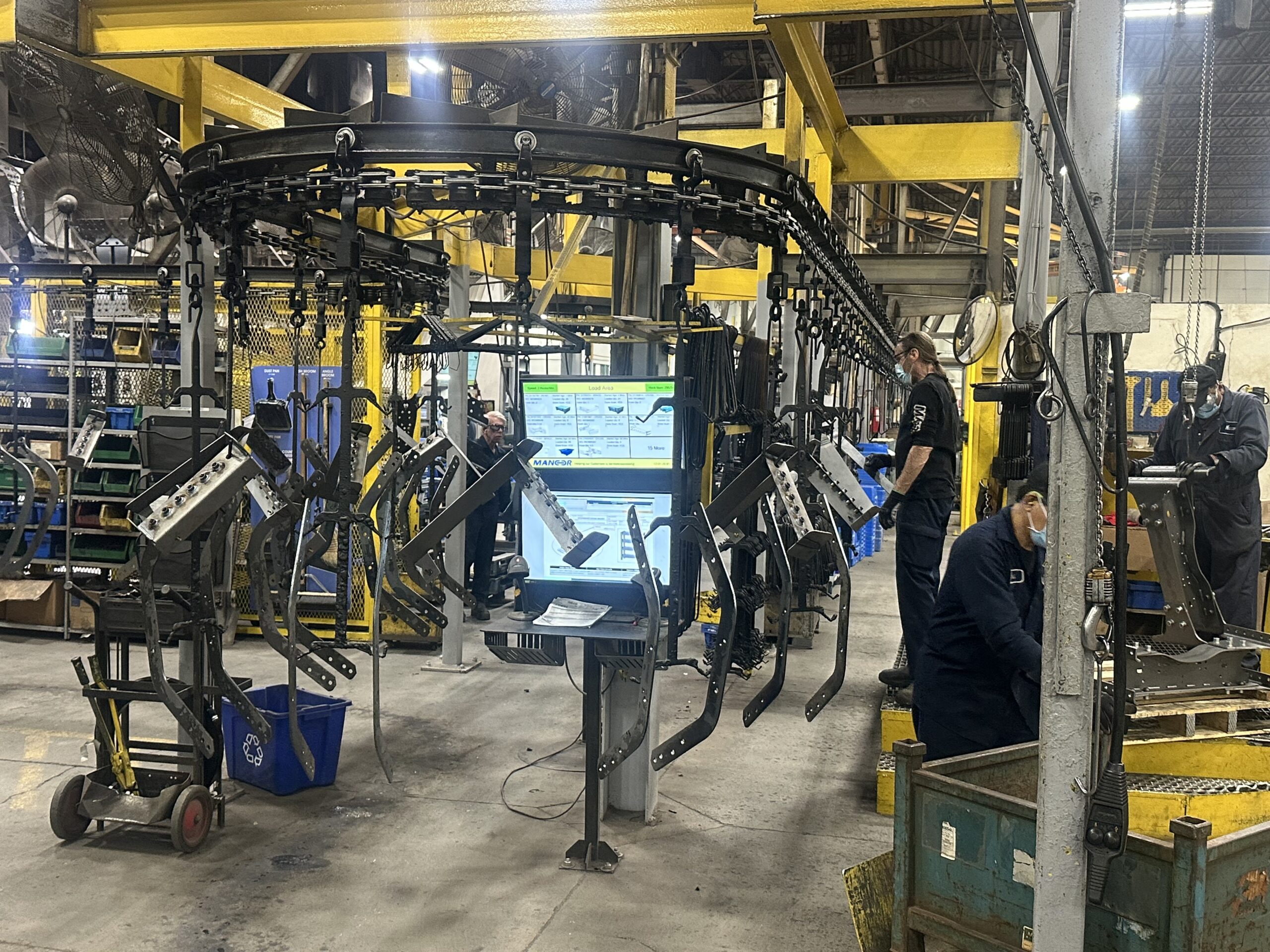 Operators working alongside a powder coating conveyor line with a job display screen on the shop floor