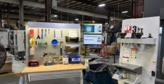 A CNC machining workstation on a shop floor with tools organized on a pegboard and a monitor displaying quality management for manufacturers software in the background.