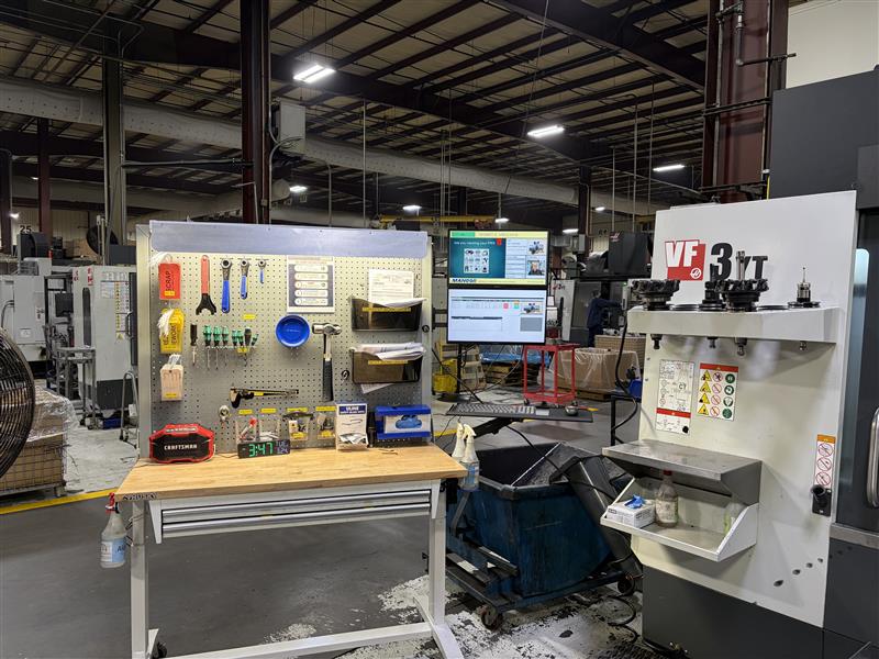 A CNC machining workstation on a shop floor with tools organized on a pegboard and a monitor displaying quality management for manufacturers software in the background.