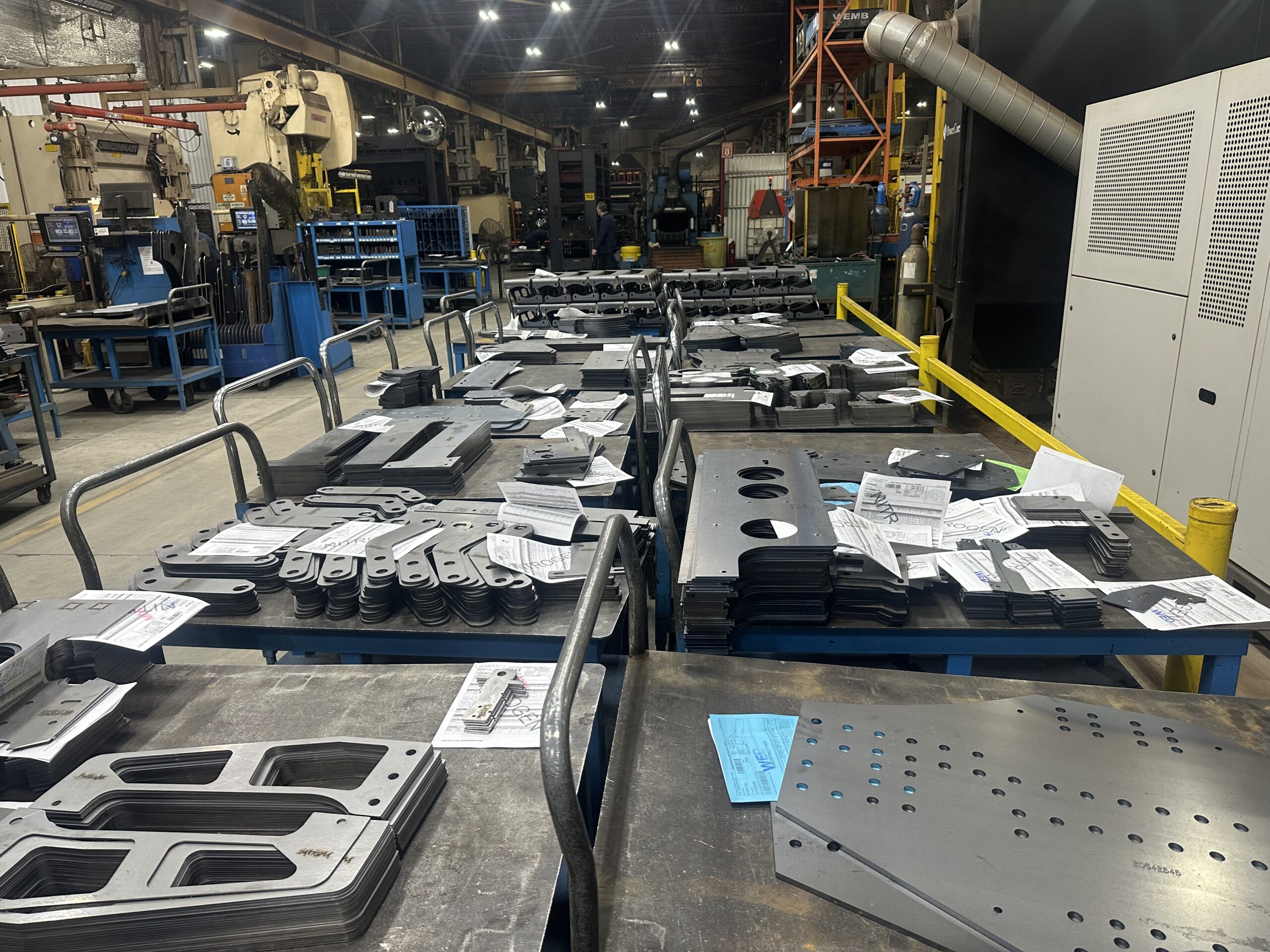 fabrication ERP features: cut sheet metal parts staged on work tables with paper job travelers in a fabrication shop
