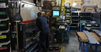 Operator at a brake press on a fabrication shop floor, with line-side monitor and job status indicators visible in the background.