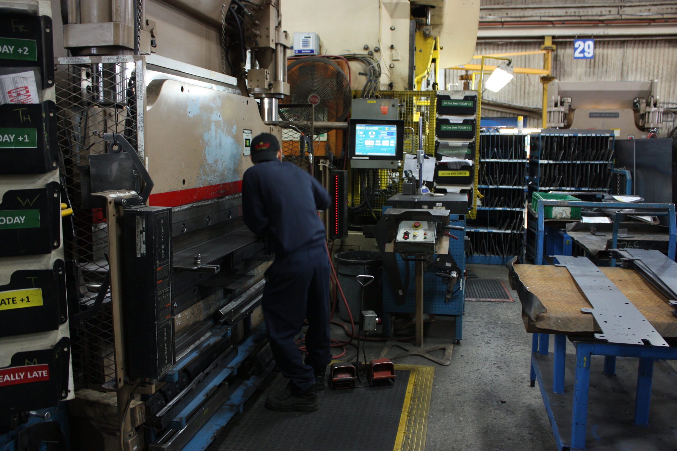 Operator at a brake press on a fabrication shop floor, with line-side monitor and job status indicators visible in the background.