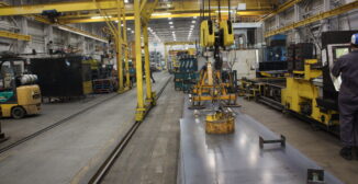 Steel fabrication shop floor with overhead crane and raw plate material, showing a manufacturing work center where inventory accuracy and shop floor execution meet.