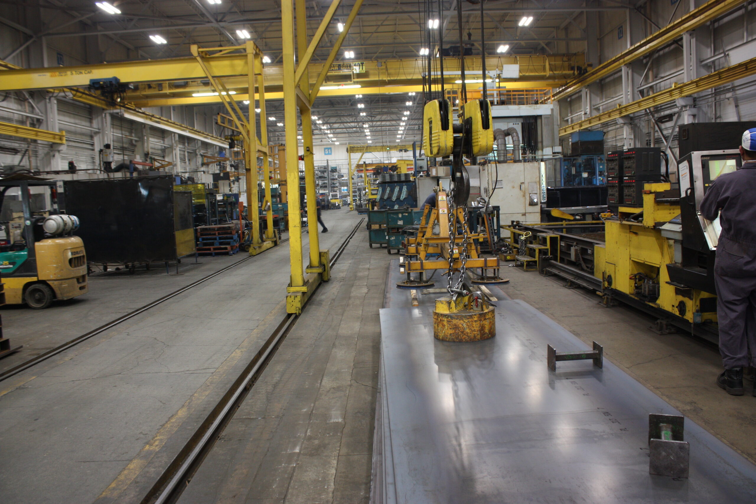 Steel fabrication shop floor with overhead crane and raw plate material, showing a manufacturing work center where inventory accuracy and shop floor execution meet.